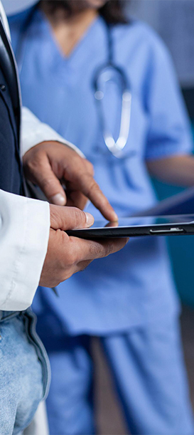 Primer plano de las manos de un profesional de la salud con bata blanca utilizando una tableta digital. En el fondo desenfocado, se aprecia a otro miembro del equipo médico con uniforme azul y un estetoscopio. La imagen simboliza el uso de la tecnología y la digitalización de datos en el entorno clínico.
