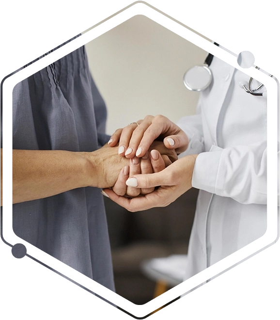 Primer plano que muestra a una profesional de la salud con bata blanca y estetoscopio, sosteniendo con ambas manos la mano de una paciente en un gesto de apoyo, consuelo y empatía. La imagen se enfoca en la conexión humana y el cuidado médico, con un fondo neutro y desenfocado.