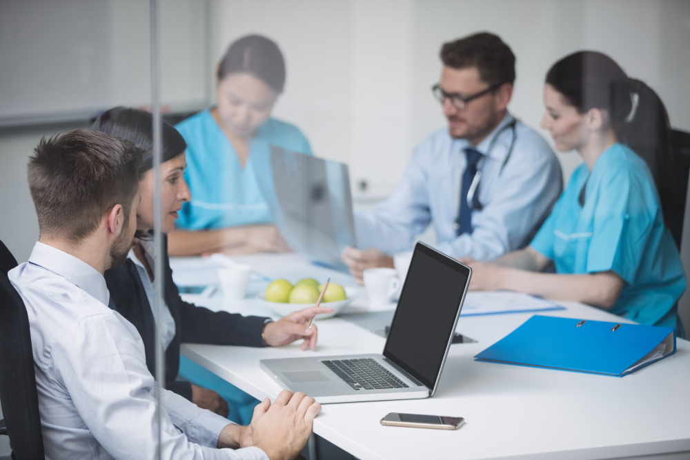 Ilustración conceptual de un entorno de trabajo tecnológico y futurista. En un amplio espacio de oficina con grandes ventanales y vista a una ciudad, se observa a un grupo de profesionales de la salud en uniformes azules analizando pantallas holográficas flotantes que muestran datos biométricos y diagramas de la anatomía humana. Una de las pantallas incluye el texto. Integración tecnológica para optimizar procesos. En el centro y a la derecha, personal con traje ejecutivo utiliza tabletas y visores de realidad virtual. El ambiente cuenta con servidores de datos con luces azules y estaciones de trabajo modernas, sugiriendo un centro de control de alta tecnología.